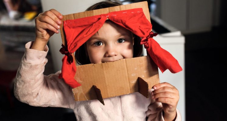 Caucasian girl child plays in a cardboard theater with a red curtain, the child himself makes toys at home, children's creativity