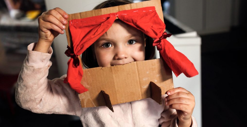 Caucasian girl child plays in a cardboard theater with a red curtain, the child himself makes toys at home, children's creativity